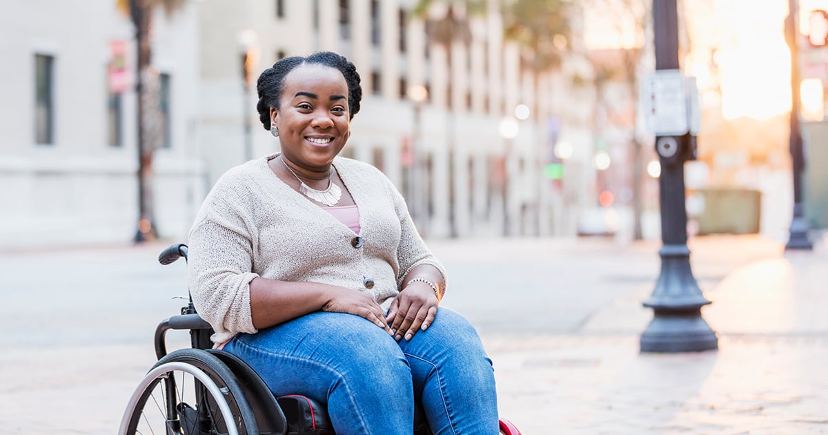 A mid adult African-American woman in her 30s in a wheelchair outdoors, in the city, smiling at the camera. She has spina bifida.