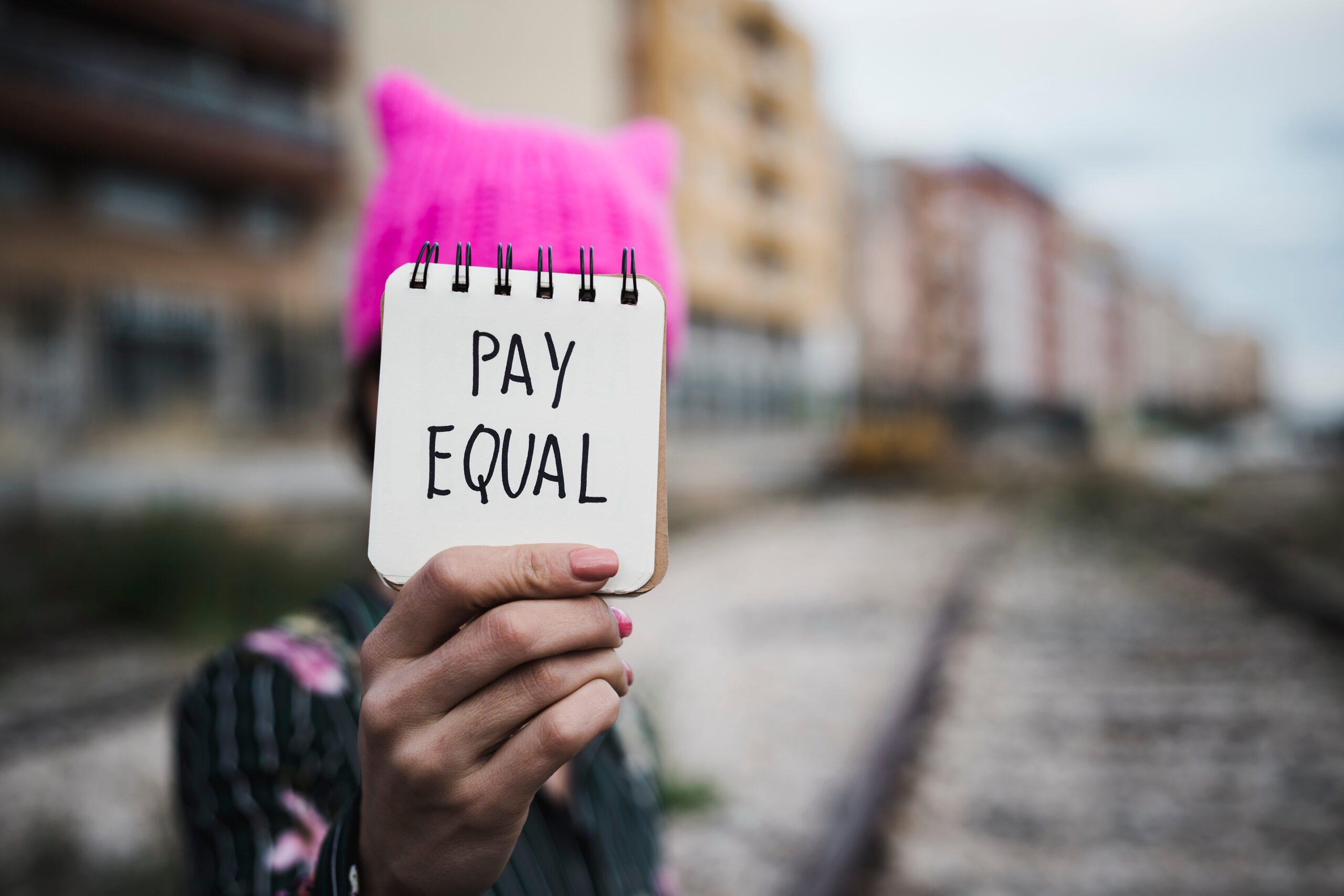 closeup of a young woman outdoors wearing a pink pussycat hat showing a piece of paper in front of her face with the text pay equal written in it