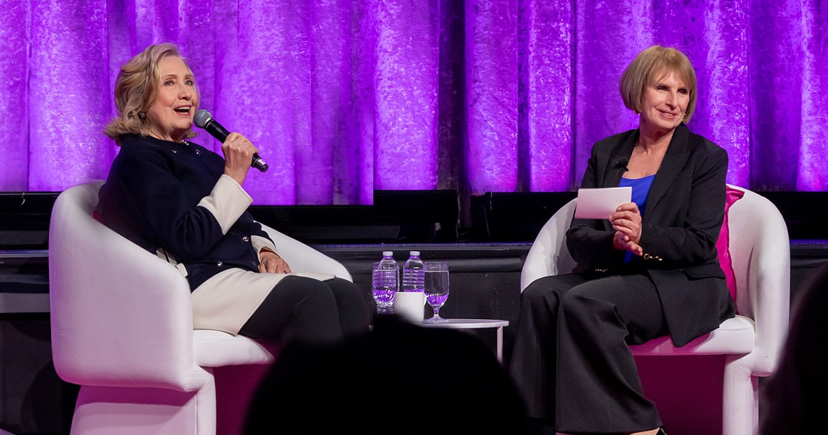 Hillary Rodham Clinton speaking into a microphone, sitting on a stage with Lorraine Hariton at the Catalyst Awards.
